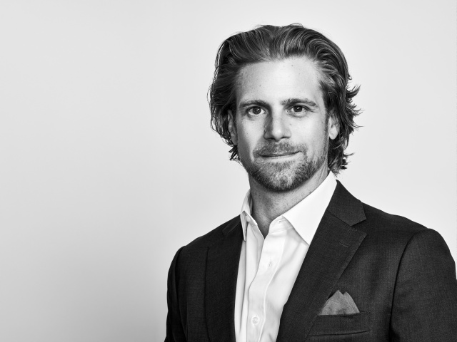 A white man with a tight beard poses on a white background in a suit with a pocket square.