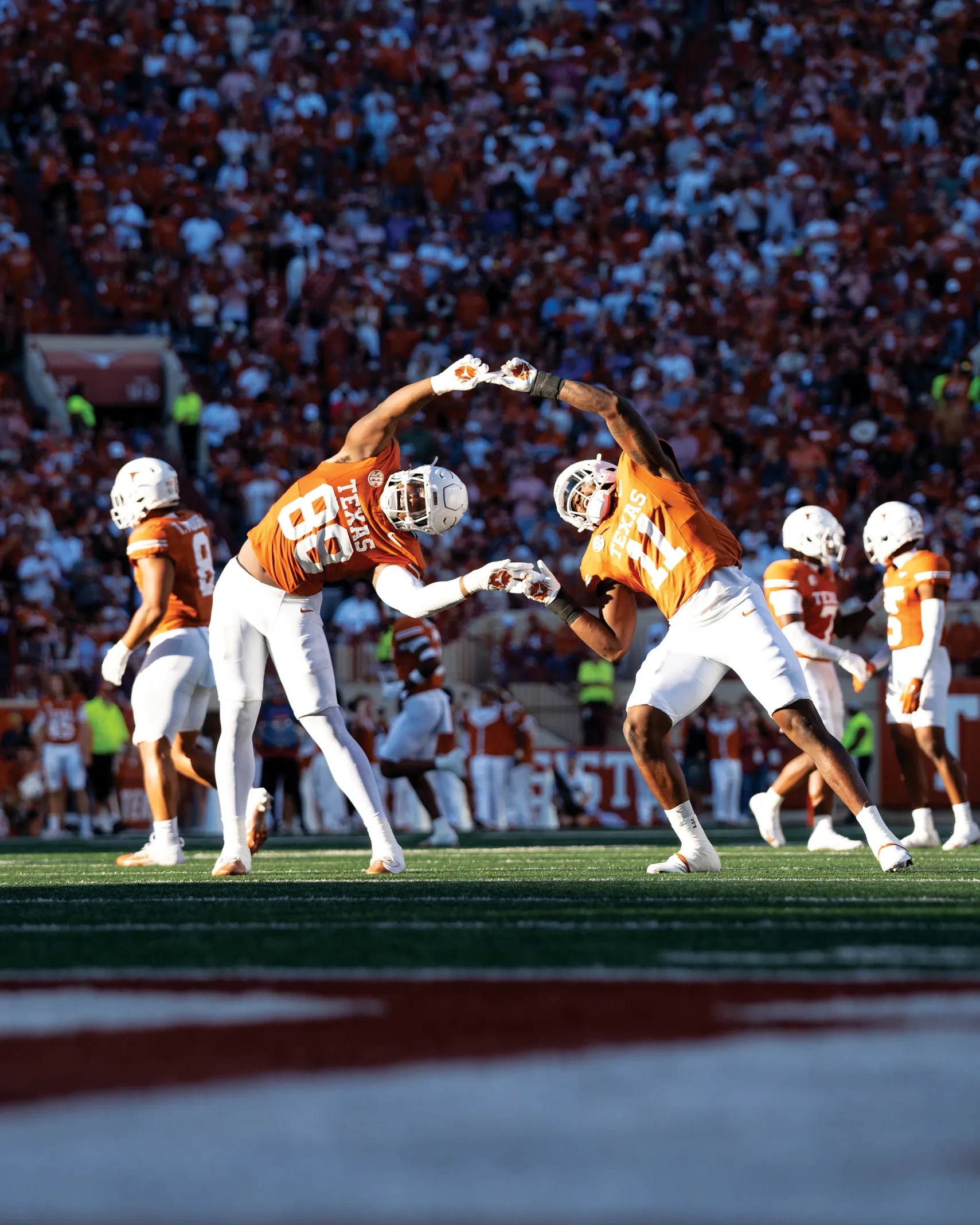 Two football players in orange celebrate on the field by forming their hands in the shape of a heart.