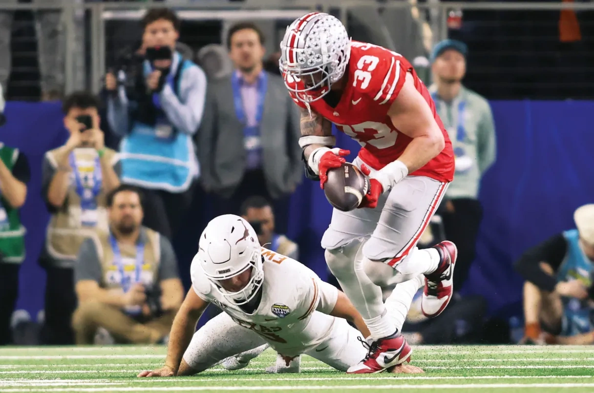 An Ohio State football player in red has a football in his hand, over a white football player down on the field.