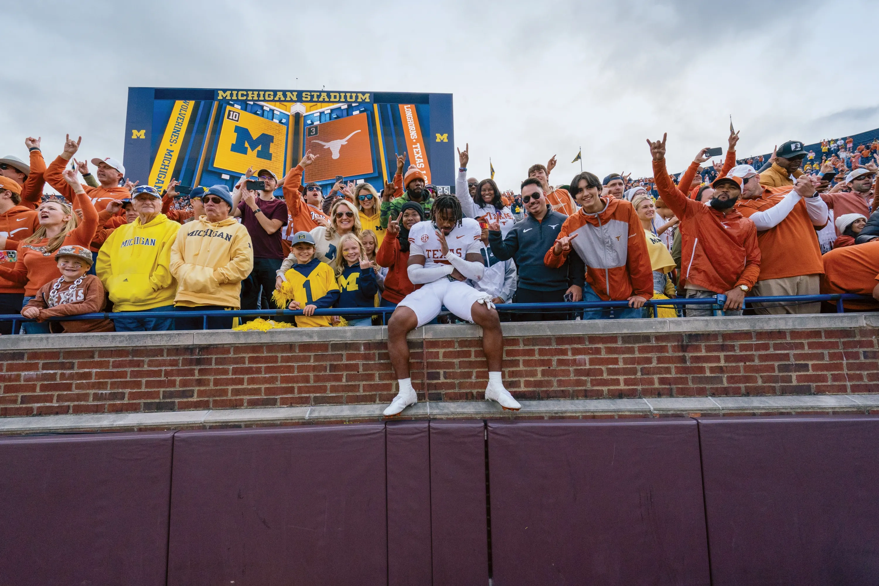 A football player in white sits in the stands of the Michigan football stadium surrounded by fans in burnt orange.